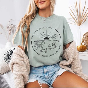 May include: A light blue t-shirt with a black graphic design of a beach scene with a sun, waves, an umbrella, and crabs. The text on the graphic says "Leaving tracks on the sand making lasting memories".