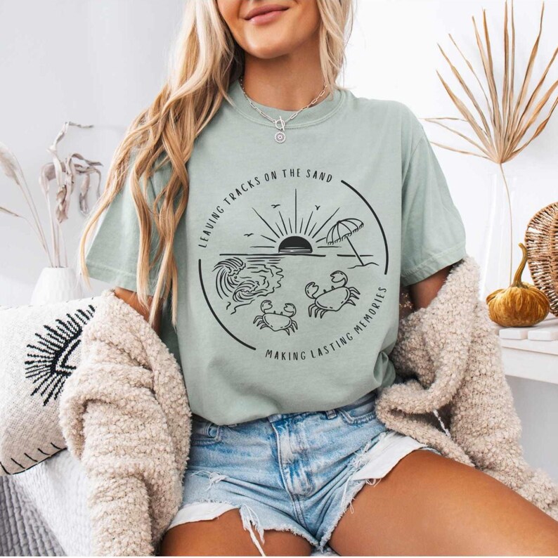 May include: A light blue t-shirt with a black graphic design of a beach scene with a sun, waves, an umbrella, and crabs. The text on the graphic says "Leaving tracks on the sand making lasting memories".