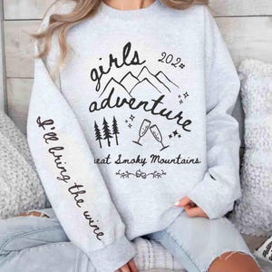 May include: A light gray sweatshirt with the words "Girls Adventure 2023" and "Great Smoky Mountains" in black script. The sleeve reads "I'll bring the wine". The design includes mountain and tree graphics.
