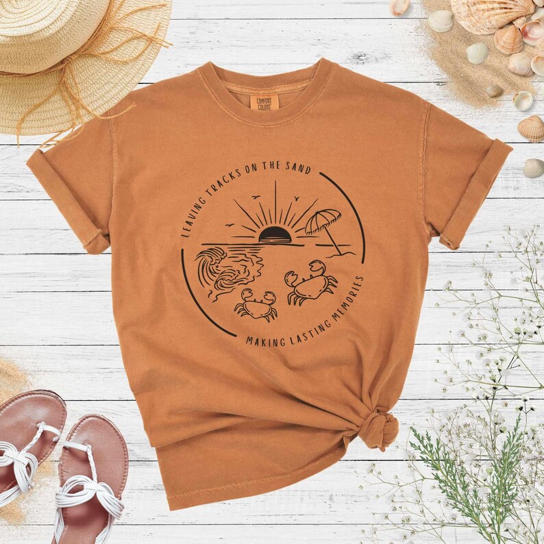 May include: A brown t-shirt with a graphic of a sunset over the ocean with two crabs and the text "Leaving tracks on the sand making lasting memories".