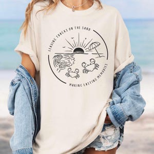 May include: A white t-shirt with a black graphic design of a beach scene with a sun, waves, an umbrella, and two crabs. The text "Leaving Tracks on the Sand Making Lasting Memories" is written around the graphic.