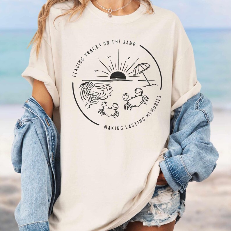 May include: A white t-shirt with a black graphic design of a beach scene with a sun, waves, an umbrella, and two crabs. The text "Leaving Tracks on the Sand Making Lasting Memories" is written around the graphic.