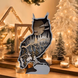 May include: A wooden owl-shaped decoration with a layered design. The owl features a detailed interior scene of trees, clouds, and a moon. The color palette includes shades of brown, gray, and black, creating a natural and rustic aesthetic. The owl is displayed on a wooden surface.