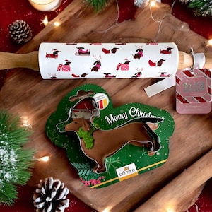 Peut inclure: Un rouleau à pâtisserie blanc avec un motif de teckels portant des bonnets de Père Noël et tenant des cadeaux. Le rouleau à pâtisserie est sur une surface en bois avec un emporte-pièce en forme de teckel vert et blanc et une étiquette cadeau rouge et blanche qui dit "The Dachshund Squad".