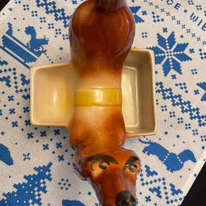 May include: A brown and white ceramic dachshund figurine with a yellow band around its middle. The figurine is holding a small, square, cream-colored dish.