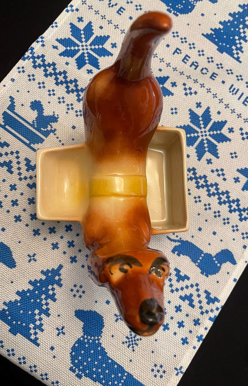 May include: A brown and white ceramic dachshund figurine with a yellow band around its middle. The figurine is holding a small, square, cream-colored dish.