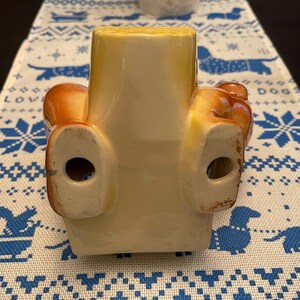 May include: A ceramic figurine of a dog sitting with its legs outstretched. The dog is brown and yellow with a white underside. The figurine has two holes in the bottom.