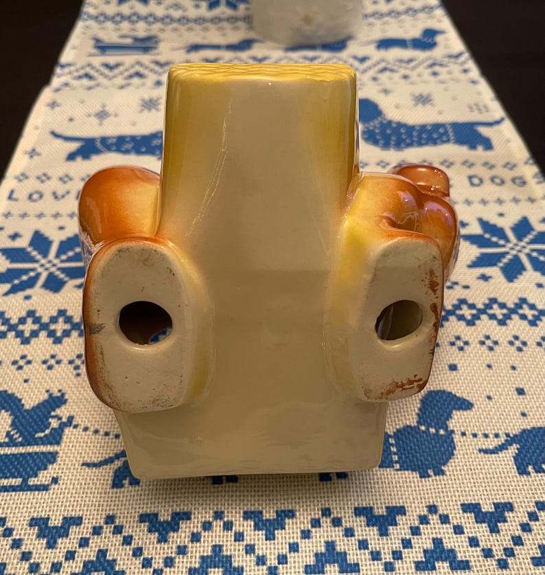 May include: A ceramic figurine of a dog sitting with its legs outstretched. The dog is brown and yellow with a white underside. The figurine has two holes in the bottom.