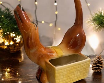 May include: A brown ceramic dachshund planter with a yellow wicker basket inside. The dachshund is standing on its hind legs with its head tilted back and its tail curled up.