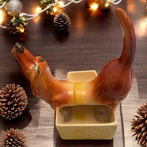 May include: A ceramic dachshund figurine with a brown and tan glaze. The dachshund is curled up in a sitting position with its tail curled over its back. The figurine has a small planter in the center of its back.