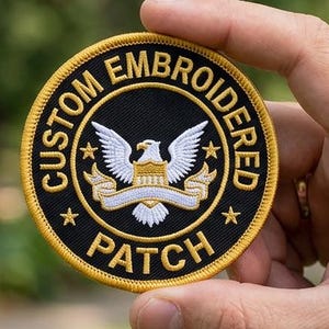 May include: A circular embroidered patch with a black center and a gold border. The text CUSTOM EMBROIDERED PATCH is stitched in gold around the edge. A white eagle with spread wings sits above a white banner in the center, flanked by two gold stars.