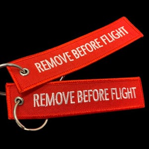 May include: Two red rectangular keychains with the text "REMOVE BEFORE FLIGHT" embroidered in white. Each keychain has a silver metal ring. The background is black.