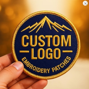 May include: Round navy blue embroidered patch with gold text and border. The patch features a mountain graphic above the words "CUSTOM LOGO" and "EMBROIDERY PATCHES". The patch is held by a hand.