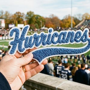 Könnte beinhalten: Ein blauer Chenille-Aufnäher mit weißem Rand, der das Wort Hurricanes in einer Schreibschrift zeigt. Der Aufnäher hat eine strukturierte, flauschige Oberfläche und wird vor einem unscharfen Hintergrund eines Fußballstadions gehalten.