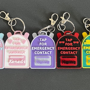 May include: Five colorful backpack-shaped keychains with silver key rings. Each keychain has the text "TAP FOR EMERGENCY CONTACT" and a space for a name. Colors include pink, purple, black, and yellow.