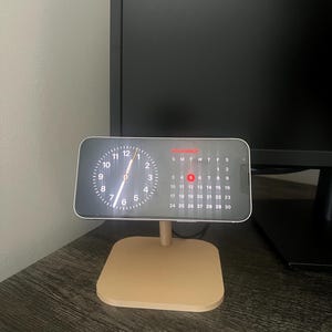 May include: A gold metal stand with a round top and three legs holds a black smartphone with a clock face and a calendar showing the month of November. The stand is on a dark wood surface.