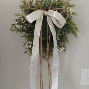 Peut inclure: Décoration murale ornée d'une base en conifère vert, de petites baies blanches et d'un grand nœud en ruban de satin blanc et en ficelle naturelle. De longs rubans blancs pendent. Idéale pour une décoration saisonnière ou toute l'année.