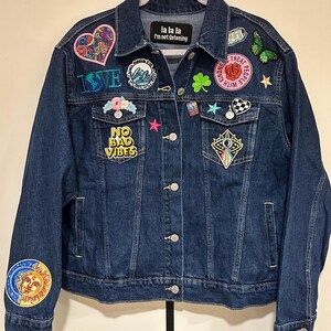 May include: A dark blue denim jacket with a variety of colorful patches, including a heart, a wave, a clover, a star, a checkered flag, and a sun and moon. The text "la la la I'm not listening" is on a black patch on the jacket.