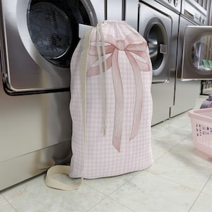 May include: Pink and white gingham laundry bag with a large pink bow. The bag is sitting in front of a washing machine.
