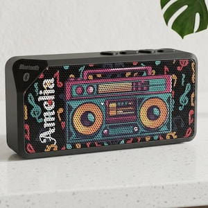 Puede incluir: Altavoz Bluetooth negro con un colorido diseño de boombox retro. El altavoz tiene el nombre "Amelia" impreso en el lateral. Notas musicales y claves de sol decoran la parte frontal de malla del altavoz.