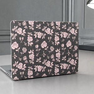 May include: Laptop computer with a floral design case. The case has a dark grey background with a pattern of pink roses and green leaves. The laptop is on a light grey desk, with a silver desk lamp in the background.