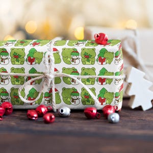 May include: A white gift box wrapped in green wrapping paper with a pattern of frogs wearing Santa hats and holding holly. The box is tied with twine and has red jingle bells around it.