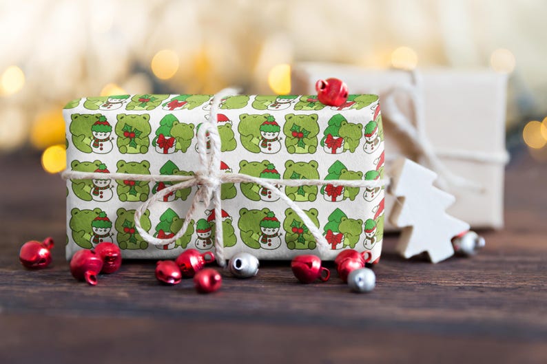 May include: A white gift box wrapped in green wrapping paper with a pattern of frogs wearing Santa hats and holding holly. The box is tied with twine and has red jingle bells around it.