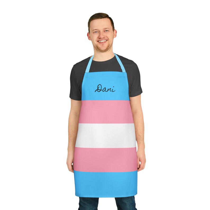 May include: A kitchen apron featuring horizontal stripes in light blue, pink, and white, mimicking the transgender pride flag. The apron includes a neck loop and waist ties, with the name Dani written in black cursive script on the top blue stripe.