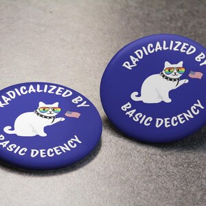 K&ouml;nnte beinhalten: Zwei blaue runde Buttons mit wei&szlig;em Text und einer wei&szlig;en Katzenillustration. Der Text lautet "RADIKALISIERT DURCH GRUNDLEGENDEN ANSTAND". Die Katze tr&auml;gt eine Regenbogen-Sonnenbrille und h&auml;lt eine kleine amerikanische Flagge.