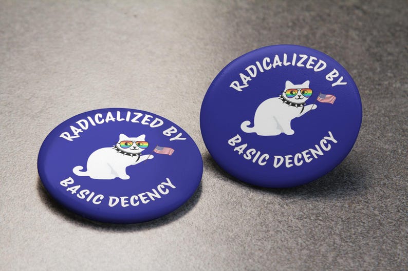 K&ouml;nnte beinhalten: Zwei blaue runde Buttons mit wei&szlig;em Text und einer wei&szlig;en Katzenillustration. Der Text lautet "RADIKALISIERT DURCH GRUNDLEGENDEN ANSTAND". Die Katze tr&auml;gt eine Regenbogen-Sonnenbrille und h&auml;lt eine kleine amerikanische Flagge.