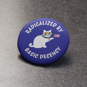 K&ouml;nnte beinhalten: Ein runder, blauer Button mit wei&szlig;em Text, der lautet: &bdquo;RADIKALISIERT DURCH GRUNDLEGENDEN ANSTAND&ldquo;. Eine wei&szlig;e Katze mit Regenbogen-Sonnenbrille und Nietenkragen h&auml;lt eine kleine amerikanische Flagge.