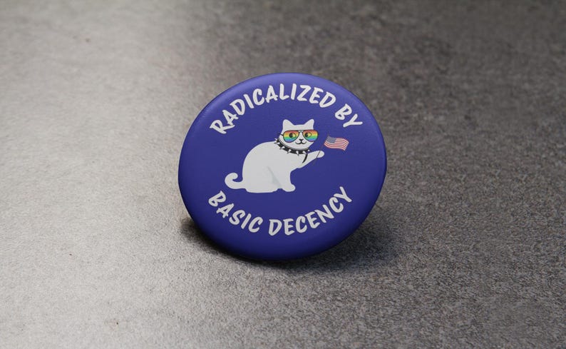 K&ouml;nnte beinhalten: Ein runder, blauer Button mit wei&szlig;em Text, der lautet: &bdquo;RADIKALISIERT DURCH GRUNDLEGENDEN ANSTAND&ldquo;. Eine wei&szlig;e Katze mit Regenbogen-Sonnenbrille und Nietenkragen h&auml;lt eine kleine amerikanische Flagge.
