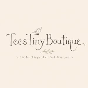 May include: A logo design on a cream background featuring the dark brown text TeesTinyBoutique in a stylized serif font. A small bird sits on the letter T with a tiny heart above it. Below the text is a small leaf sprig and the phrase little things that feel like you.