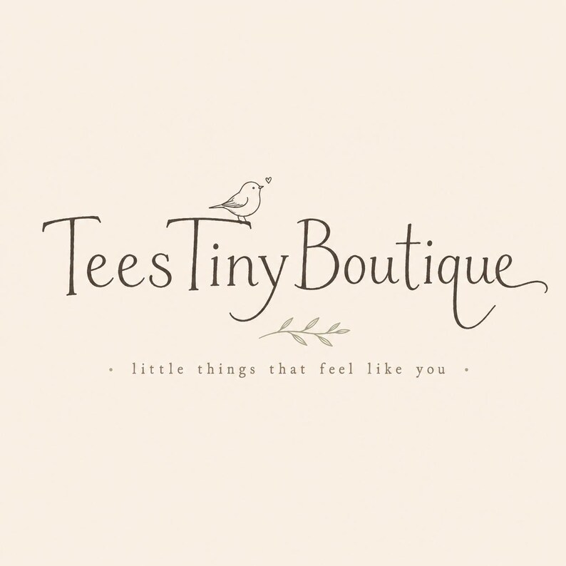 May include: A logo design on a cream background featuring the dark brown text TeesTinyBoutique in a stylized serif font. A small bird sits on the letter T with a tiny heart above it. Below the text is a small leaf sprig and the phrase little things that feel like you.
