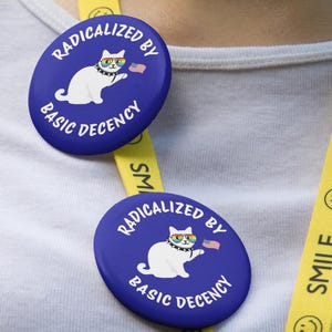 K&ouml;nnte beinhalten: Zwei blaue ovale Buttons mit dem Text "RADIKALISIERT DURCH GRUNDANSTAND" und einer Cartoon-Katze mit Regenbogenbrille und einer kleinen amerikanischen Flagge. Ein gelbes Schl&uuml;sselband mit dem Wort "SMILE" und einem Smiley ist sichtbar.