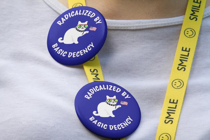 K&ouml;nnte beinhalten: Zwei blaue ovale Buttons mit dem Text "RADIKALISIERT DURCH GRUNDANSTAND" und einer Cartoon-Katze mit Regenbogenbrille und einer kleinen amerikanischen Flagge. Ein gelbes Schl&uuml;sselband mit dem Wort "SMILE" und einem Smiley ist sichtbar.