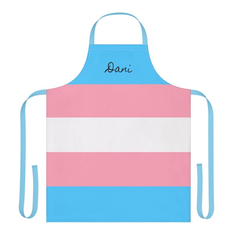 May include: A light blue apron featuring the horizontal striped pattern of the transgender pride flag in light blue, pink, and white. The upper section is light blue with the name Dani written in black cursive script centered on the chest area.