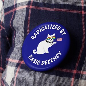 K&ouml;nnte beinhalten: Ein blauer, ovaler Button mit dem wei&szlig;en Text "RADICALIZED BY BASIC DECENCY". Der Button zeigt eine wei&szlig;e Katze mit Regenbogen-Sonnenbrille und einem Nietenhalsband, die eine kleine amerikanische Flagge h&auml;lt.