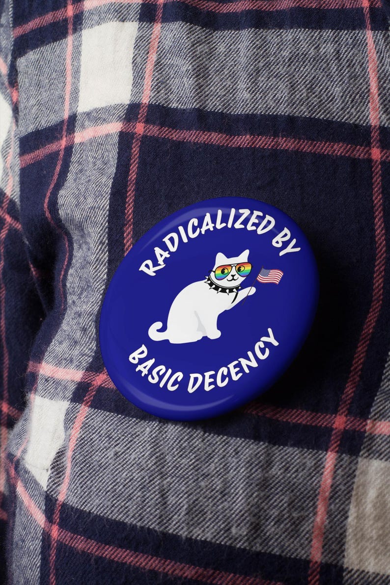 K&ouml;nnte beinhalten: Ein blauer, ovaler Button mit dem wei&szlig;en Text "RADICALIZED BY BASIC DECENCY". Der Button zeigt eine wei&szlig;e Katze mit Regenbogen-Sonnenbrille und einem Nietenhalsband, die eine kleine amerikanische Flagge h&auml;lt.