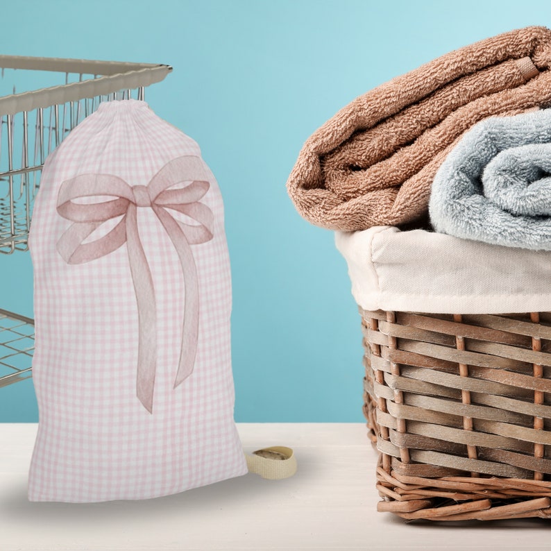 以下が含まれることがあります： ピンクと白のギンガムチェックのランドリーバッグに大きな茶色のリボンが付いている。バッグは白い表面に置かれ、茶色のタオルが入った籐製のランドリーバスケットの隣にある。