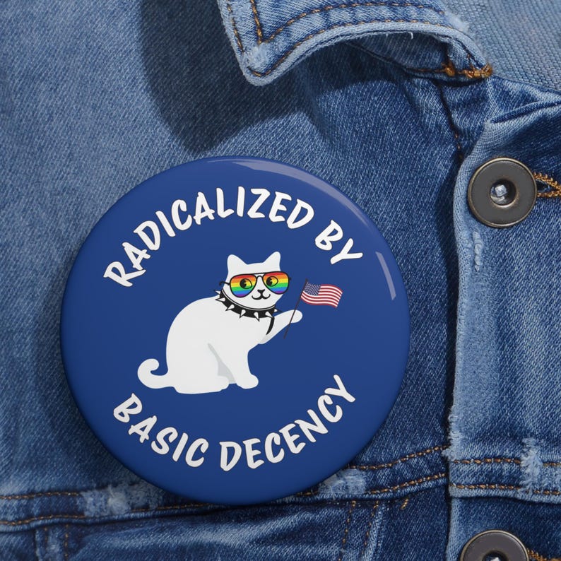 May include: A round, blue button with the text "RADICALIZED BY BASIC DECENCY" encircling a white cat wearing rainbow sunglasses and a studded collar, holding a small American flag. The button is set against a denim background.