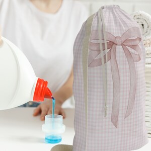 以下が含まれることがあります： ピンクと白のギンガムチェックのランドリーバッグに大きなピンクのリボンが付いています。バッグは柔らかく丈夫な生地で作られており、巾着式になっています。洗濯洗剤のボトルが計量カップに注がれています。