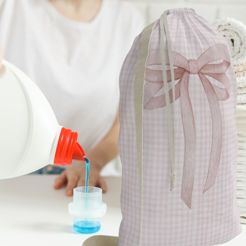 以下が含まれることがあります： ピンクと白のギンガムチェックのランドリーバッグに大きなピンクのリボンが付いています。バッグは柔らかく丈夫な生地で作られており、巾着式になっています。洗濯洗剤のボトルが計量カップに注がれています。