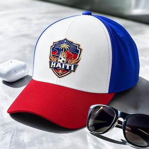 May include: A baseball cap with a red brim, white front panel, and blue top and back. The cap features a shield-shaped emblem with the word "HAITI" and a design including a palm tree and soccer ball.