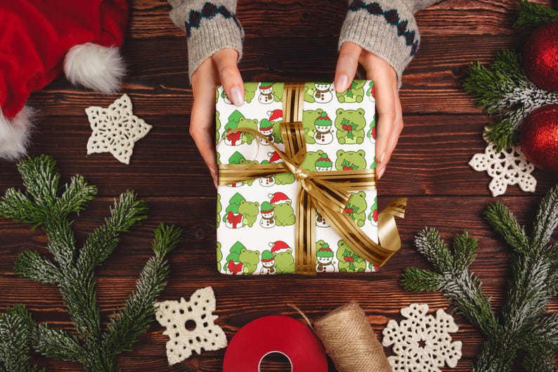 May include: A white gift box wrapped in green and white paper with a repeating pattern of frogs wearing Santa hats and holding Christmas trees. The box is tied with a gold ribbon.