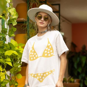Puede incluir: Una camiseta blanca con un diseño de bikini de lunares amarillos. La camiseta se usa con un sombrero blanco y gafas de sol. El diseño incluye un top de bikini y una braguita de bikini.