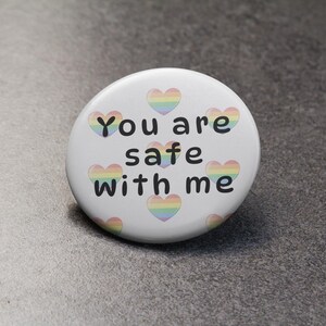 Puede incluir: Un botón blanco con el texto "You are safe with me" en negro. El botón presenta diseños de corazones de colores arcoíris. El botón tiene forma redonda y es probablemente un pin o insignia.