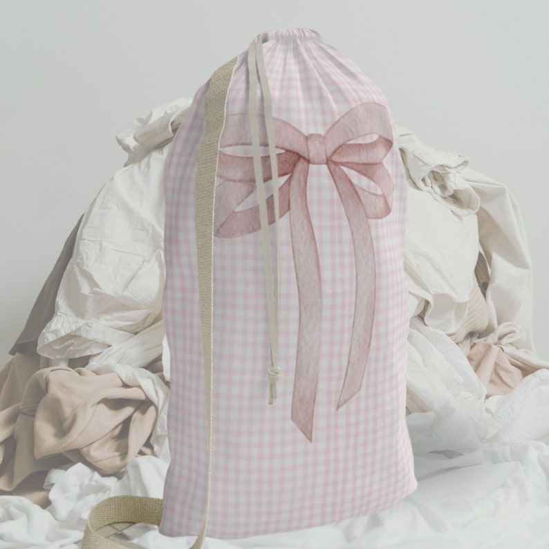 以下が含まれることがあります： ピンクと白のギンガムチェックのランドリーバッグで、大きなピンクのリボンが付いています。バッグは巾着式で、ベージュの綿ひもが付いています。