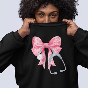 Könnte beinhalten: Schwarzes Sweatshirt mit einem rosafarbenen Band und einem Stethoskop-Design. Das Band hat ein Blumenmuster.