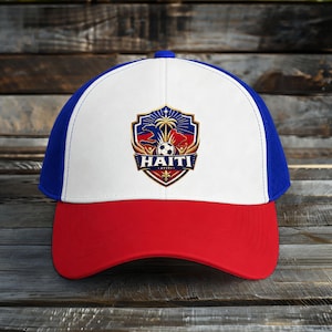 May include: A baseball cap with a red brim, white front panel, and blue mesh back. The front features a shield-shaped emblem with the word "HAITI" and a soccer ball. The emblem also includes a palm tree and the words "AVITI."